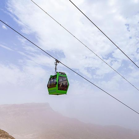 עיין סוחנה Tolip Resort El Galala Majestic מראה חיצוני תמונה The photo shows a green cable car suspended in the air, traveling along overhead cables. The backdrop features a hazy landscape, likely mountainous or elevated terrain, with a cloudy sky. The scene suggests a scenic transportation method, possibly us