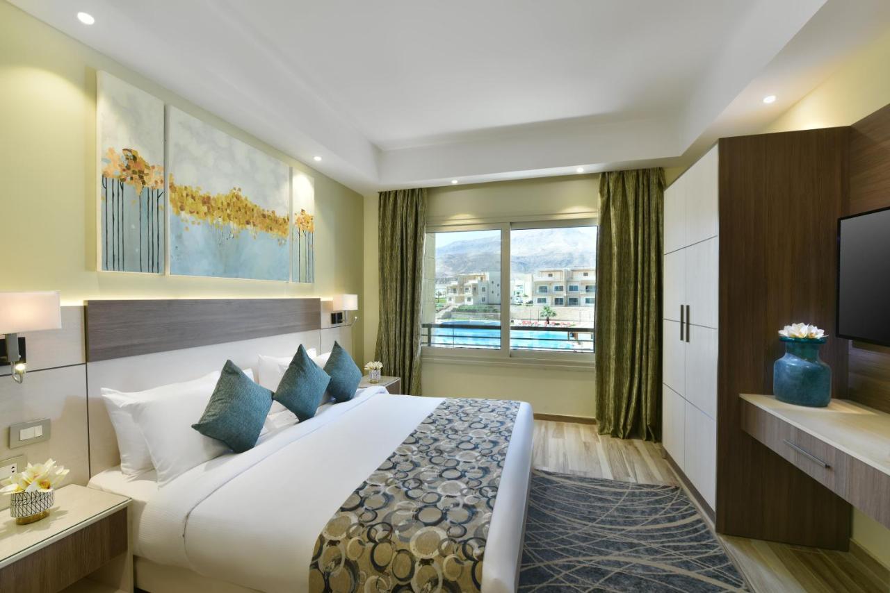 עיין סוחנה Tolip Resort El Galala Majestic מראה חיצוני תמונה The photo shows a modern hotel room. It features a large bed with white bedding and several decorative pillows in shades of teal. Above the bed, there are two pieces of wall art with a landscape theme. The room has a large window with green curtains,