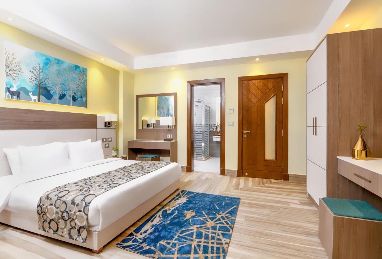 עיין סוחנה Tolip Resort El Galala Majestic מראה חיצוני תמונה The photo shows a well-decorated hotel room. There is a large bed with white linens and a decorative throw blanket. The room features a wooden wardrobe and a bedside table with lamps. A colorful rug is placed on the floor, adding warmth to the space.