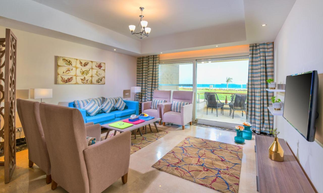 עיין סוחנה Tolip Resort El Galala Majestic מראה חיצוני תמונה The photo shows a bright and modern living room. It features a blue sofa with decorative cushions, two light-colored armchairs, and a coffee table. A large area rug adds warmth to the space, which is decorated with a subtle pattern. The walls have so