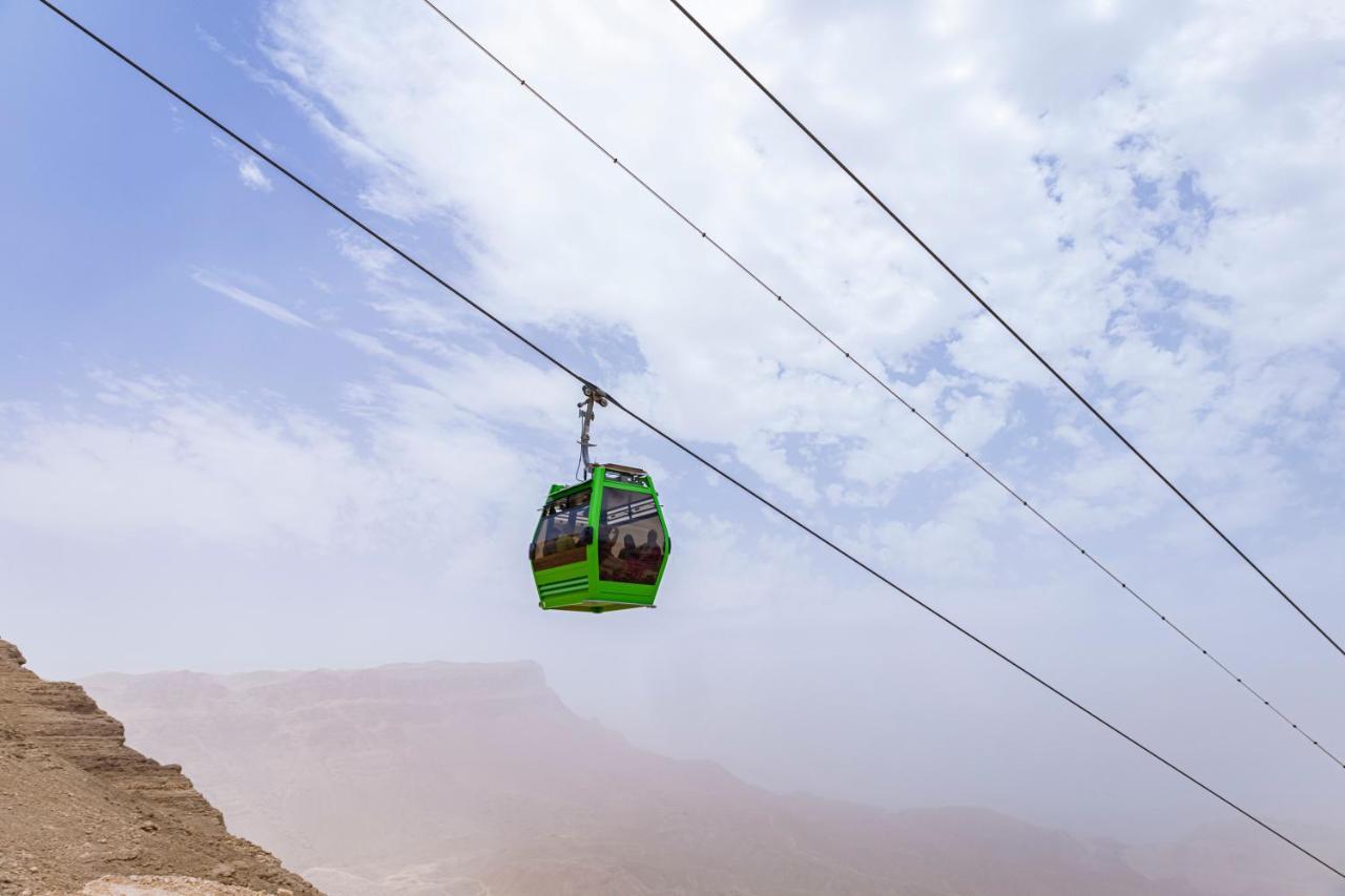 עיין סוחנה Tolip Resort El Galala Majestic מראה חיצוני תמונה The photo shows a green cable car suspended in the air, traveling along overhead cables. The backdrop features a hazy landscape, likely mountainous or elevated terrain, with a cloudy sky. The scene suggests a scenic transportation method, possibly us