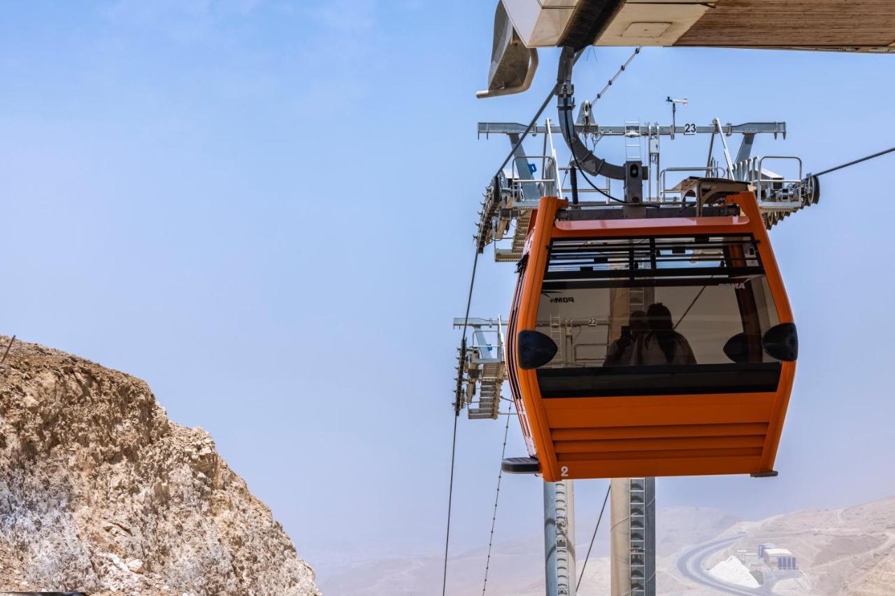 עיין סוחנה Tolip Resort El Galala Majestic מראה חיצוני תמונה The photo shows an orange gondola or cable car hanging on a cable system. The gondola appears to be suspended in the air above a rocky landscape, with a clear sky in the background. There are supporting cables and structures visible, indicating that 