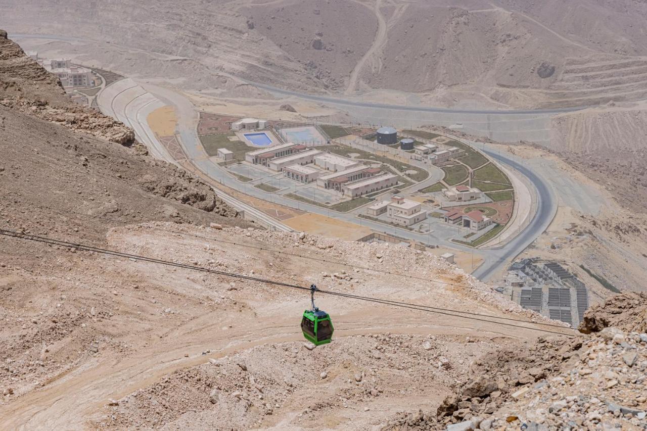 עיין סוחנה Tolip Resort El Galala Majestic מראה חיצוני תמונה The photo shows a mountainous landscape with a steep, dry terrain. In the foreground, there is a green cable car or gondola traveling along a cable. Below, a complex of buildings and roads can be seen, likely part of a resort or recreational area. Th
