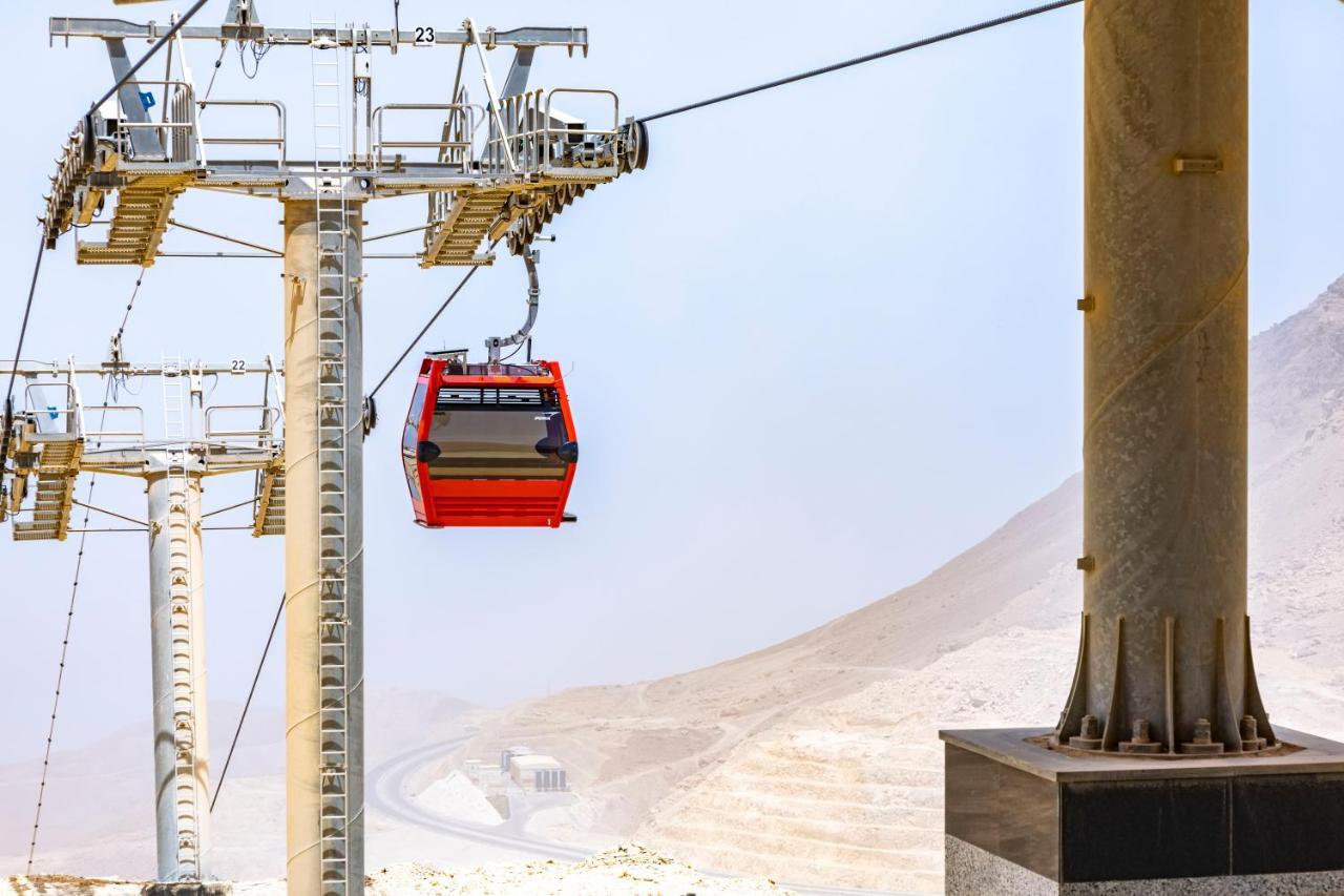 עיין סוחנה Tolip Resort El Galala Majestic מראה חיצוני תמונה The photo shows a cable car system with a single red gondola suspended mid-air. It appears to be part of a larger cable car infrastructure, with metal pylons supporting the cables above. The background features a hazy landscape, likely characterized 