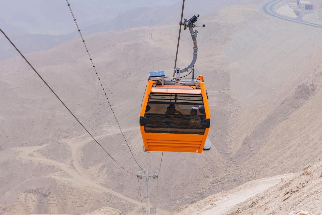 עיין סוחנה Tolip Resort El Galala Majestic מראה חיצוני תמונה The photo shows an orange cable car suspended in the air, traveling along a set of cables. It appears to be located in a mountainous or hilly area, with rugged terrain and dry, rocky landscapes visible below. The background features slopes that seem 