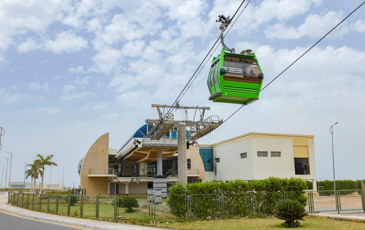 עיין סוחנה Tolip Resort El Galala Majestic מראה חיצוני תמונה The photo shows a cable car or gondola system in operation, featuring a green-colored gondola. It is suspended from cables and moving towards a station, which appears to be a modern building with a unique architectural design. The station has large w
