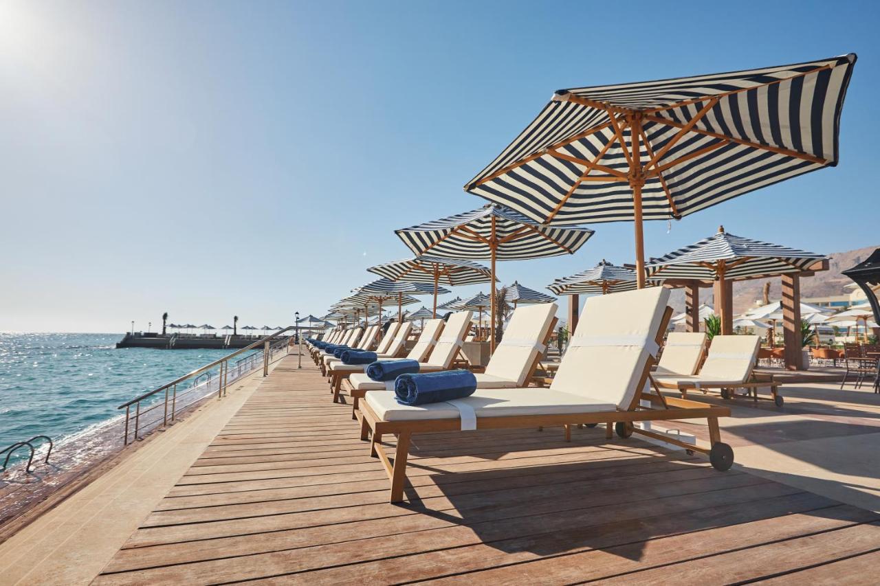 עיין סוחנה Tolip Resort El Galala Majestic מראה חיצוני תמונה The photo depicts a serene beachside setting featuring a row of sun loungers arranged on a wooden deck. Each lounger is equipped with a folded towel and shaded by striped umbrellas. In the background, the water sparkles under bright sunlight, and the