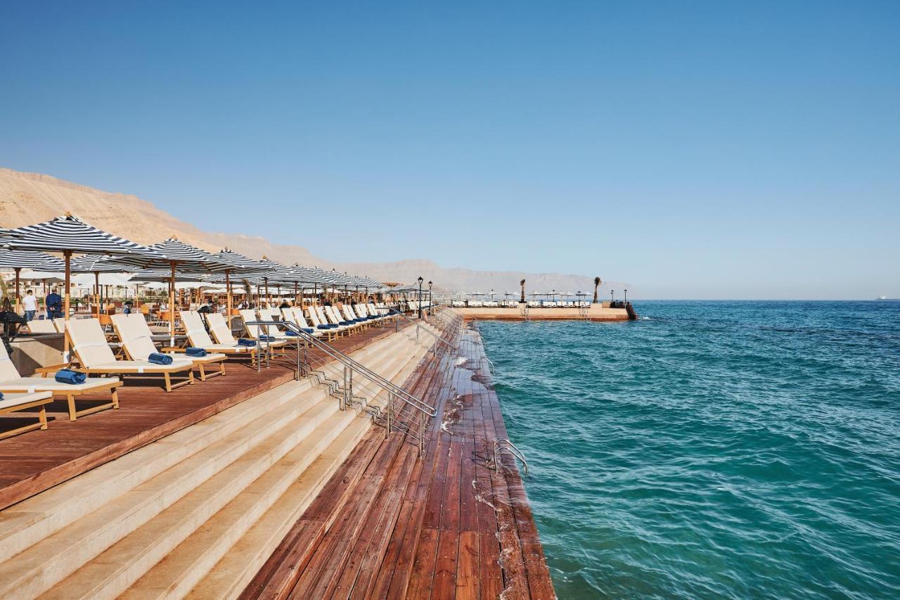עיין סוחנה Tolip Resort El Galala Majestic מראה חיצוני תמונה The photo shows a seaside area featuring a wooden deck that extends into the water. On the left side, there are several lounge chairs arranged neatly under shade structures, suggesting a relaxing atmosphere. The ocean is visible in the foreground, wi