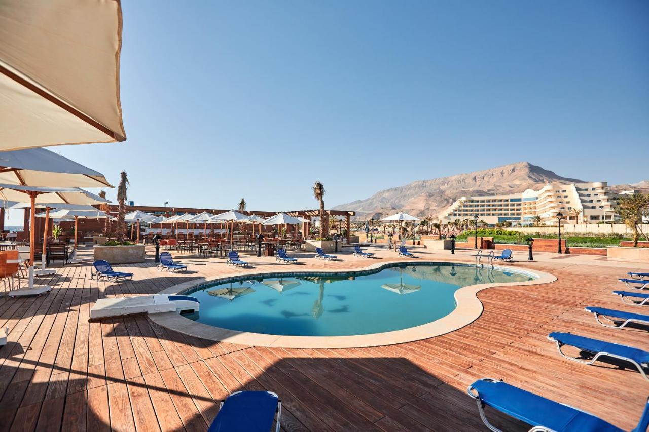 עיין סוחנה Tolip Resort El Galala Majestic מראה חיצוני תמונה The photo shows a swimming pool area surrounded by sun loungers and umbrellas. The pool has a unique, curvy shape and is situated on a wooden deck. In the background, there are several buildings, likely part of a resort or hotel, and a mountainous la