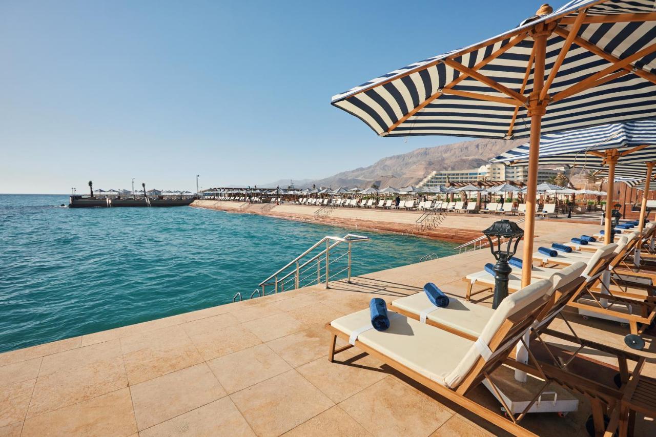 עיין סוחנה Tolip Resort El Galala Majestic מראה חיצוני תמונה The photo shows a scenic beachside area with clear blue water. In the foreground, there are several sun loungers set up on a tiled surface, with blue towels neatly rolled on each one. The loungers are shaded by striped umbrellas. In the distance, a w