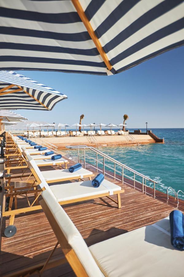 עיין סוחנה Tolip Resort El Galala Majestic מראה חיצוני תמונה The photo depicts a serene outdoor area by the waterfront. There are several lounge chairs arranged in a row, each equipped with blue towels. Large, striped umbrellas provide shade over some of the chairs. In the background, there is a wooden deck th