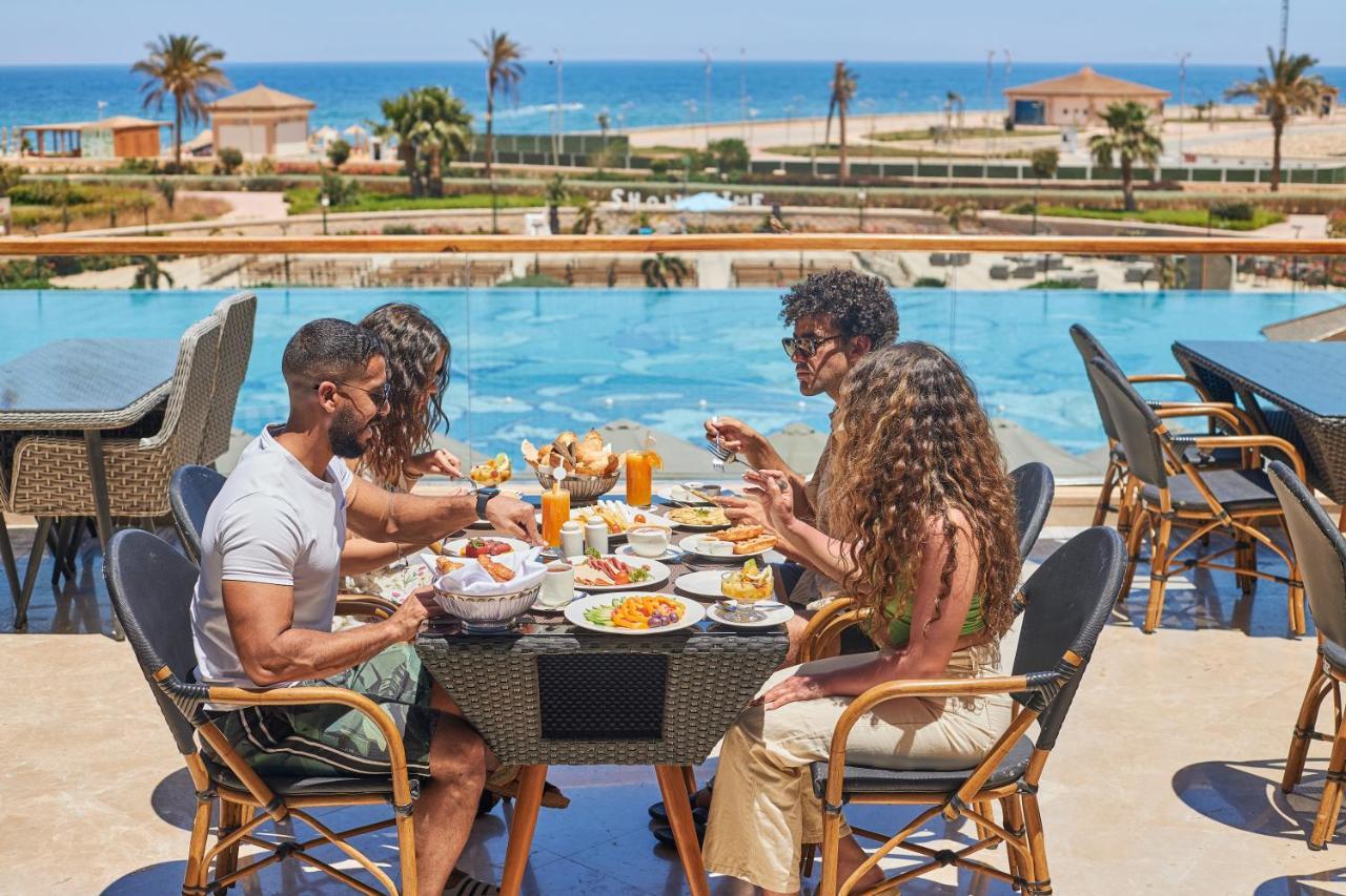 עיין סוחנה Tolip Resort El Galala Majestic מראה חיצוני תמונה The photo shows a group of five people enjoying a meal together outdoors, likely at a restaurant or cafe. They are seated at a table with a beautiful view of a swimming pool and the sea in the background. The table is filled with various dishes, and 