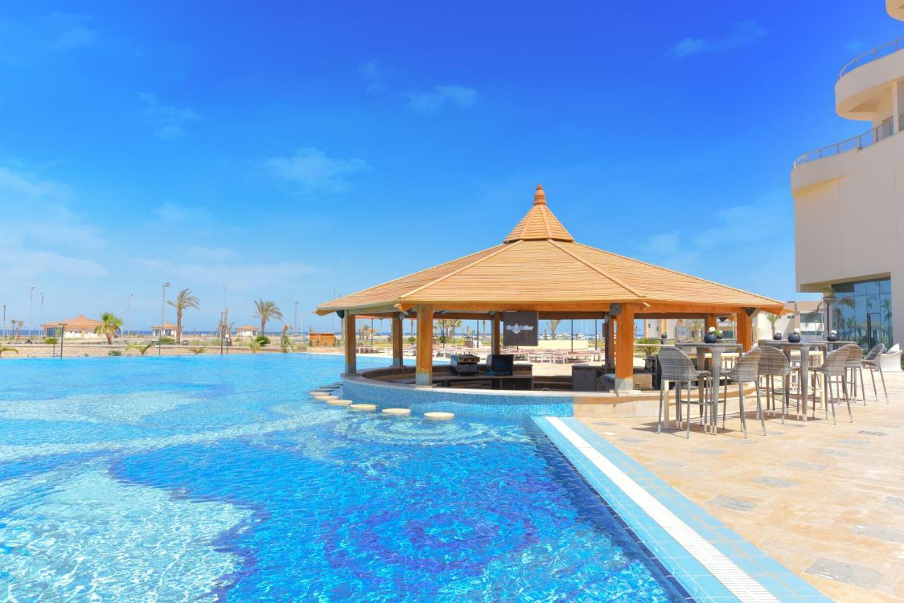 עיין סוחנה Tolip Resort El Galala Majestic מראה חיצוני תמונה The photo shows a beautiful outdoor setting featuring a large swimming pool with clear blue water. In the center, there is a thatched-roof gazebo, which serves as a shaded seating area. Surrounding the gazebo are lounge chairs and a bar area with hig