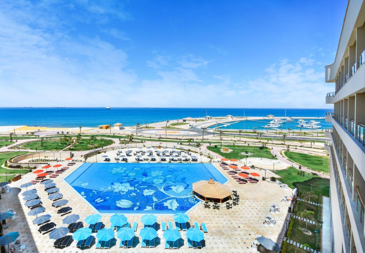 עיין סוחנה Tolip Resort El Galala Majestic מראה חיצוני תמונה The photo shows a beautiful outdoor view of a resort area featuring a large swimming pool with a decorative underwater design. Surrounding the pool are numerous sun loungers and umbrellas in a variety of colors. In the background, there is a scenic c