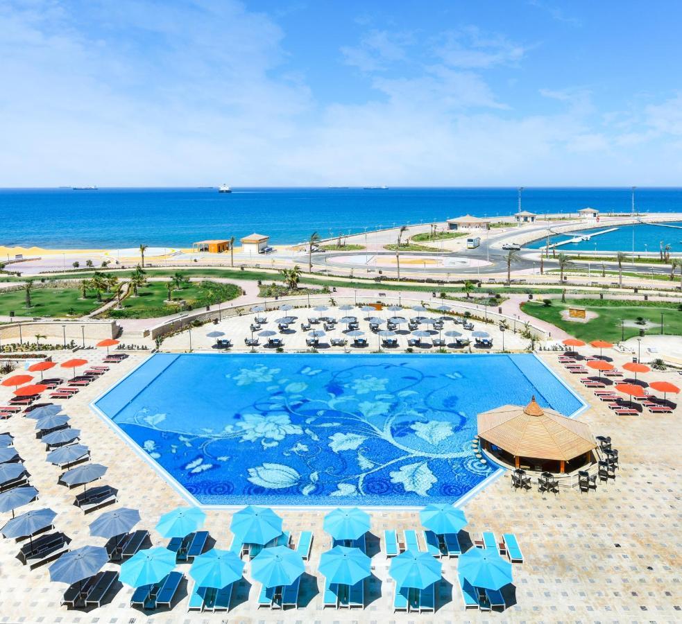 עיין סוחנה Tolip Resort El Galala Majestic מראה חיצוני תמונה The photo features a large swimming pool with a decorative design on its surface, surrounded by sun loungers and umbrellas. In the background, there is a view of the sea, with a clear blue sky and a few scattered clouds. The area is well-maintained, 
