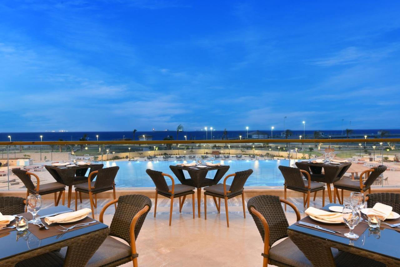 עיין סוחנה Tolip Resort El Galala Majestic מראה חיצוני תמונה The photo shows an outdoor dining area with several neatly arranged tables and chairs. The tables are elegantly set with cutlery and plates, suggesting a fine dining experience. In the background, there is a large swimming pool that reflects the blue
