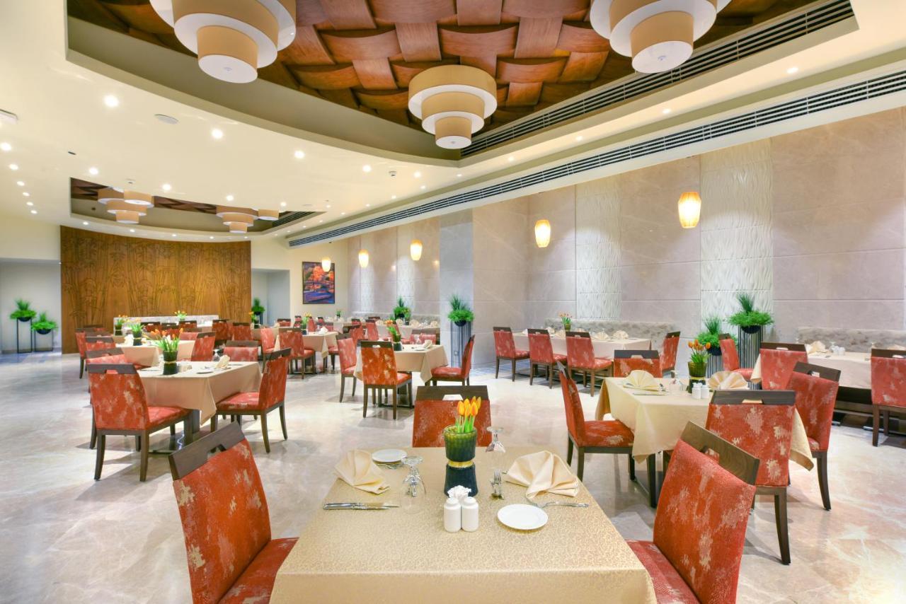 עיין סוחנה Tolip Resort El Galala Majestic מראה חיצוני תמונה The photo shows the interior of a restaurant. The space features several tables set with light-colored tablecloths and chairs with reddish upholstery. Each table is adorned with decorative napkins and has small plants in the center. The room has a mo