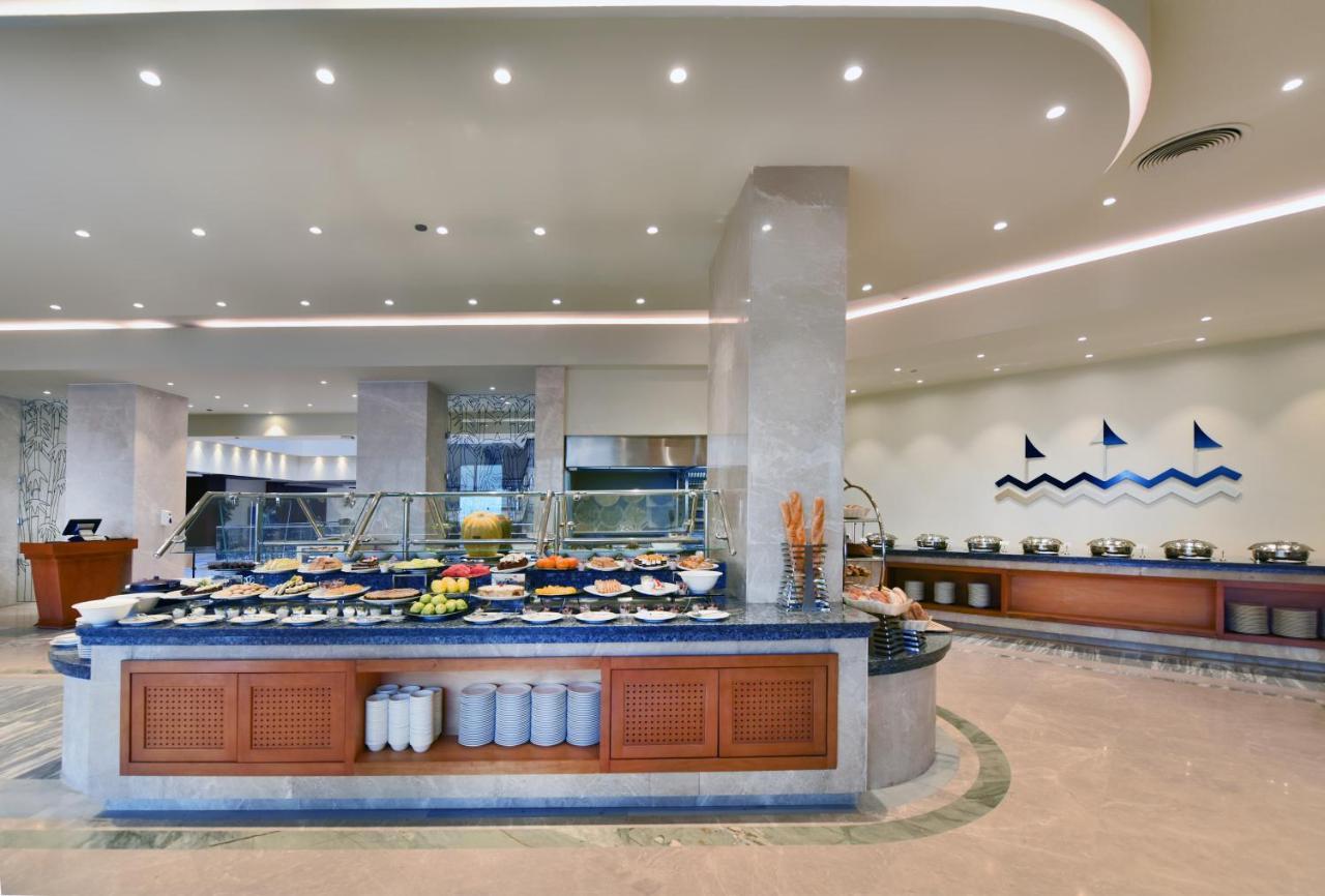 עיין סוחנה Tolip Resort El Galala Majestic מראה חיצוני תמונה The image shows a buffet-style dining area in a restaurant or hotel. There are several food stations with a variety of dishes displayed, including fruits, salads, and other food items. The layout features a large, central island with a wooden base an