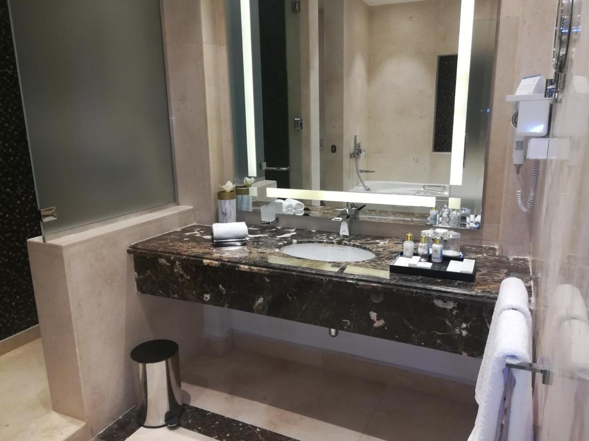 עיין סוחנה Tolip Resort El Galala Majestic מראה חיצוני תמונה The photo shows a modern bathroom with elegant design features. There is a large vanity made of dark marble with a sink and a set of toiletries neatly arranged on top. A mirror with built-in lighting is mounted above the sink. To the side, there is a