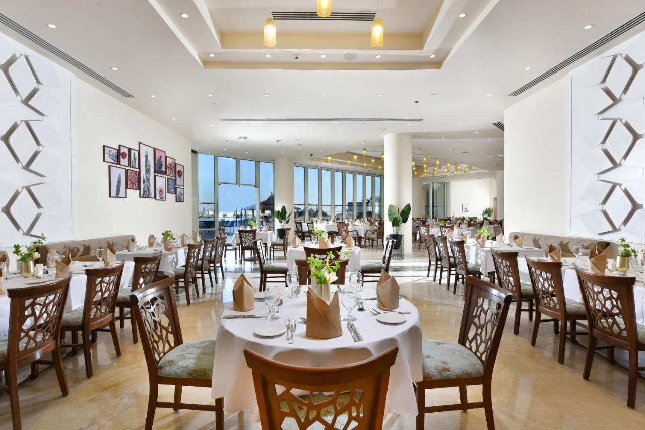 עיין סוחנה Tolip Resort El Galala Majestic מראה חיצוני תמונה The photo displays a spacious and elegantly designed restaurant interior. It features numerous tables set for dining, with white tablecloths, neatly folded napkins, and decorative centerpieces of flowers. The seating consists of wooden chairs that co