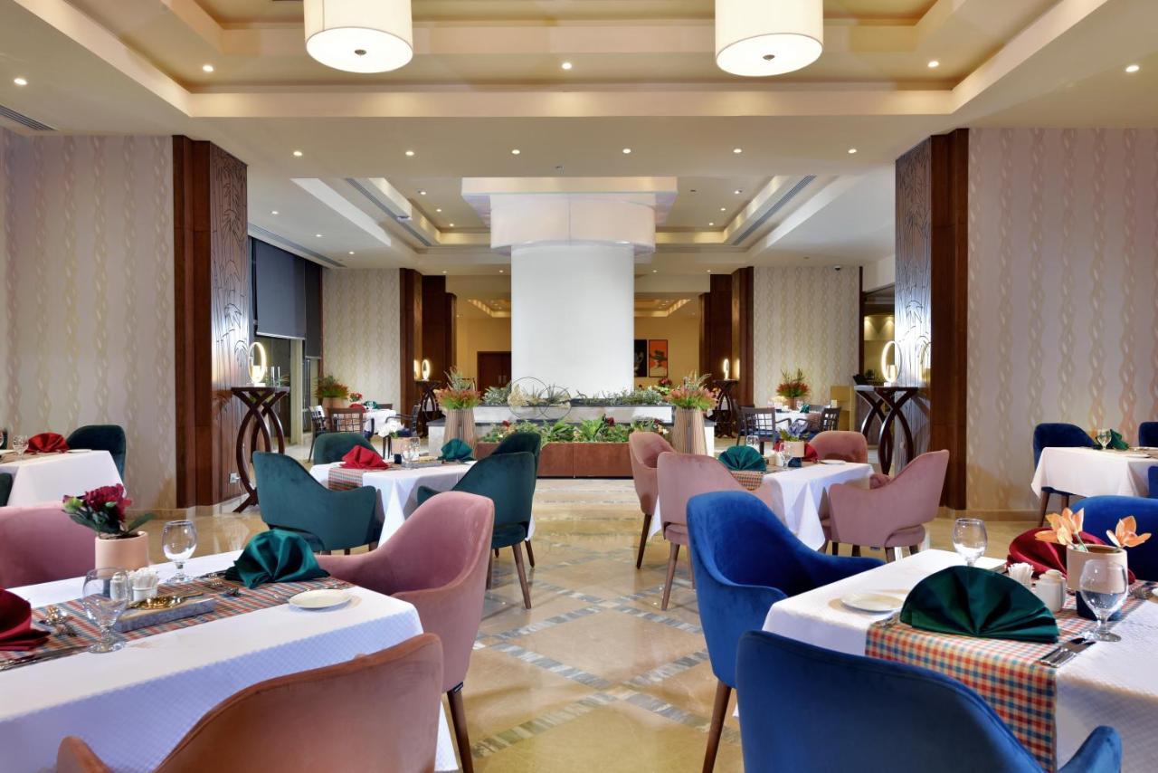 עיין סוחנה Tolip Resort El Galala Majestic מראה חיצוני תמונה The photo shows a spacious and elegantly designed restaurant interior. The layout includes several round tables dressed in white tablecloths, each set with decorative plates, cutlery, and colorful napkins. The chairs are upholstered in a variety of c